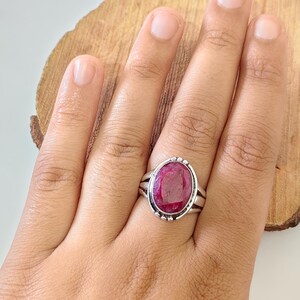 May include: A silver ring featuring a large, oval-shaped, deep pinkish-red gemstone set in a polished metal bezel. The band is constructed with a split-shank design, and the setting is accented with small, decorative silver beads along the outer edge.