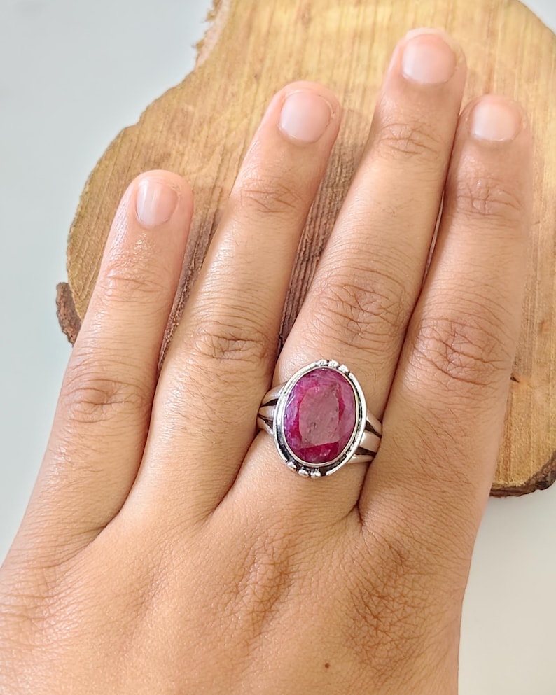 May include: A silver ring featuring a large, oval-shaped, deep pinkish-red gemstone set in a polished metal bezel. The band is constructed with a split-shank design, and the setting is accented with small, decorative silver beads along the outer edge.
