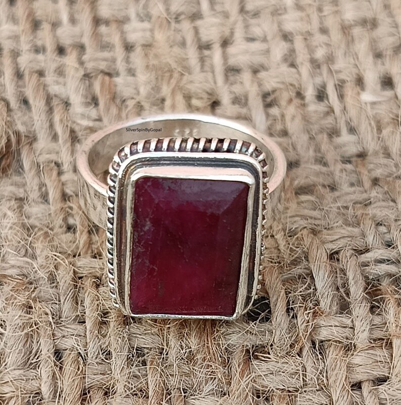 May include: A silver ring featuring a rectangular red gemstone set within a silver bezel. The ring has a textured band and is engraved with the text "SilverSpinByGopal".