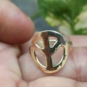 May include: A gold-coloured ring with a peace symbol design. The ring has a hammered texture and is set against a blurred background. The peace sign is a prominent, open design.