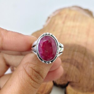 May include: A silver ring featuring a large, oval-shaped, deep red gemstone set in a polished metal bezel. The setting is accented with small, decorative silver beads around the perimeter of the stone, and the band has a split-shank design.