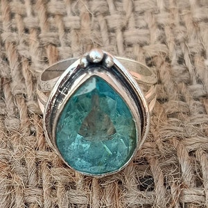 May include: A silver ring featuring a large, pear-shaped, light blue gemstone set in a silver bezel. The ring has a textured band and a decorative detail on the top.