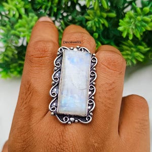 May include: A silver-toned ring with a large, rectangular moonstone. The moonstone has a milky white base with blue and iridescent flashes. The ring has an ornate, scrollwork design around the stone, and the text "DevJewellrs" is visible.