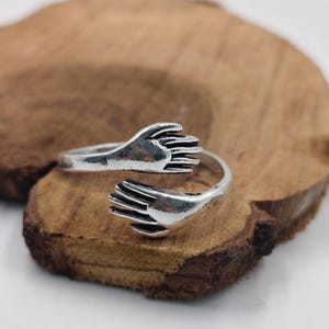 Peut inclure: Une bague argentée avec un design ouvert, représentant deux mains qui semblent s'embrasser. La bague est posée sur un fond en bois, créant un contraste entre le métal et le grain naturel du bois.