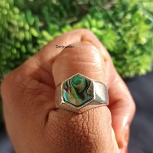 May include: A silver ring with a hexagonal abalone shell inlay. The iridescent shell features shades of green, blue, and purple. The ring is held up by a hand, with the text "Devjewellrs" visible in the background.