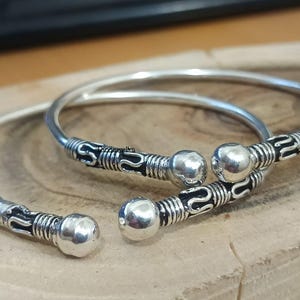 May include: Two silver-colored bangles with decorative detailing. Each bangle features a rounded, ball-shaped accent and intricate patterns. The design includes black accents, adding contrast to the silver metal. These bracelets are likely handmade.