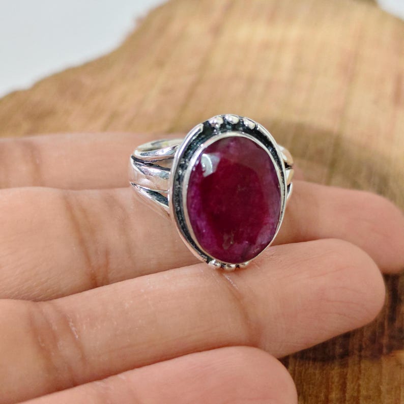 May include: A silver ring featuring a large, oval-shaped, deep red gemstone set in a polished metal bezel. The band of the ring is split into multiple strands that converge at the sides, providing a sturdy structure for the prominent central stone.