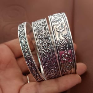 May include: Three silver bangles with floral and leaf patterns. The bangles are held in a hand, showcasing the intricate details of the engraved designs. The bangles have a polished finish, reflecting light and adding to their visual appeal. The jewelry is a classic accessory.