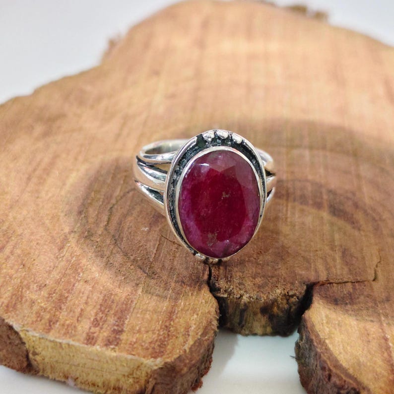 May include: A silver ring featuring a large, oval-shaped, faceted deep red gemstone set in a decorative metal bezel. The band of the ring has a split design that connects to the central setting, which is framed by small, raised silver beads.