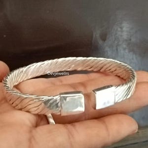 Op de afbeelding: Een zilverkleurige manchetarmband met een getextureerd, gevlochten ontwerp. De armband heeft een rechthoekige sluiting en is aan één uiteinde open. De tekst "devjewellrs" is zichtbaar.