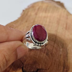 May include: A silver ring featuring a large, oval-shaped, deep red ruby gemstone set in a polished metal bezel. The band displays a split-shank design with small decorative metal spheres accenting the base of the stone setting.