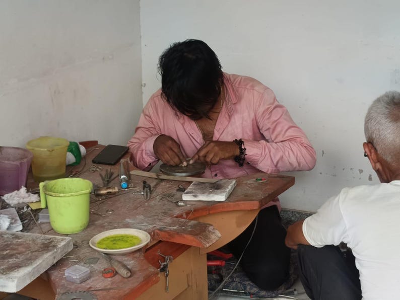 Peut inclure: Une personne portant une chemise rose travaille sur un bijou &agrave; un &eacute;tabli. Il y a divers outils et fournitures sur l'&eacute;tabli, dont un petit bol en m&eacute;tal, un seau vert et un t&eacute;l&eacute;phone portable. Une autre personne est assise en arri&egrave;re-plan.