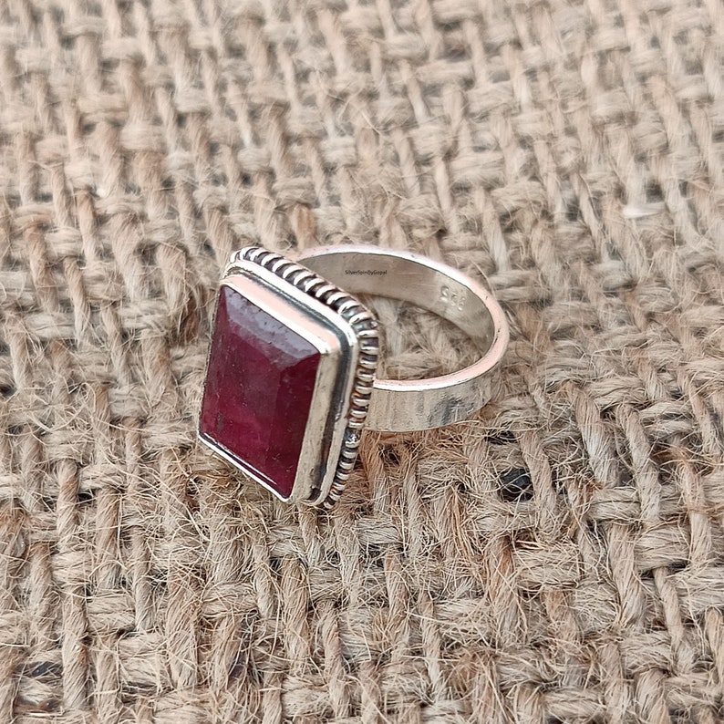 May include: A silver ring featuring a square red gemstone set in a silver bezel. The ring has a textured band and is displayed against a hessian backdrop.