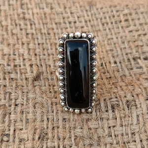 Könnte beinhalten: Ein rechteckiger Ring aus schwarzem Onyx mit einem silberfarbenen, perlenbesetzten Rand. Der Ring hat einen polierten, dunklen Stein, der in einen detaillierten, strukturierten Rahmen gefasst ist. Der Ring wird auf einer strukturierten, neutral getönten Oberfläche präsentiert.