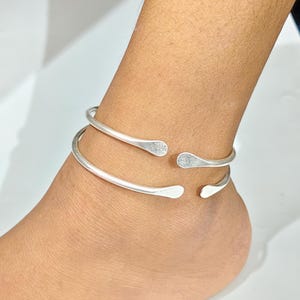 Puede incluir: Un par de tobilleras plateadas. Cada tobillera tiene un diseño sencillo y abierto con extremos en forma de lágrima. Las tobilleras se llevan en el tobillo. La joyería es de metal brillante.