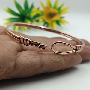 Może przedstawiać: Miedziana bransoletka z otwartym designem. Bransoletka posiada detale z owiniętego drutu na obu końcach, z jednym końcem tworzącym zapięcie na haczyk. Całość jest prosta i elegancka, odpowiednia do codziennego noszenia.