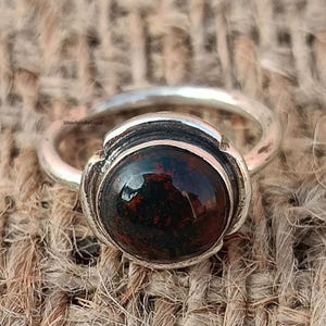 Puede incluir: Un anillo de plata con una gema grande, redonda y de color rojo oscuro. La gema tiene un aspecto moteado con inclusiones más oscuras. La banda del anillo es sencilla y el engaste alrededor de la piedra es texturizado.