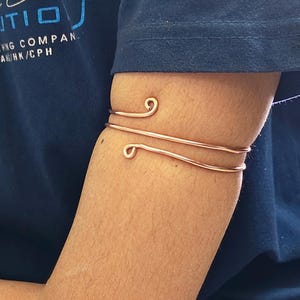 May include: Copper arm cuff with a spiral design. The cuff wraps around the arm with two parallel bands and decorative swirls at each end. The jewelry is displayed on a person's arm, against a blue shirt.