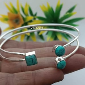 以下が含まれることがあります： ターコイズのアクセントが付いたシルバーカラーのカフブレスレット。ブレスレットには、正方形のターコイズストーンと2つの丸いターコイズストーンが付いています。ラップアラウンドデザインは、ぼやけた黄緑色の背景に対して表示されます。