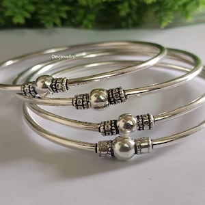 May include: Four silver bangles with decorative beaded accents. Each bangle features a smooth, rounded band with a central beaded design and end caps. The bangles are displayed on a white surface with green foliage in the background.