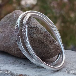 May include: Two silver hammered bangle bracelets, one slightly wider than the other, are stacked on top of each other. The bracelets are resting on a grey rock.