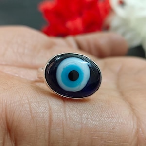 May include: Silver ring with an oval-shaped eye design. The eye has a dark blue background, a light blue iris, a white sclera, and a black pupil. The ring is set against a blurred background.