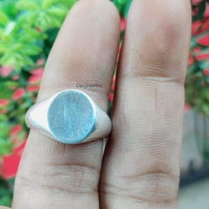 May include: A silver signet ring with a smooth, oval face. The ring is shown on a finger, with the brand name "Devjewellrs" visible. The ring's face has a light blue hue.