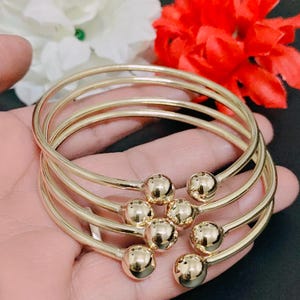 May include: Gold-colored bangles with spherical accents. The bracelets are stacked, showcasing a simple yet elegant design. The bangles are held in a hand, with red and white flowers in the background.