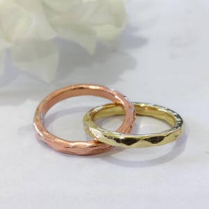 May include: Two hammered metal rings, one copper-toned and one gold-toned, interlocked. The rings have a textured surface and a simple design. The background is a soft white with a hint of a white flower.