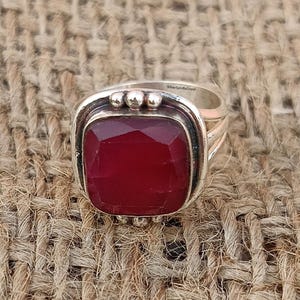 May include: A silver ring with a large, square, red gemstone set in a silver bezel. The ring has a textured band and three small silver balls on the top of the bezel.
