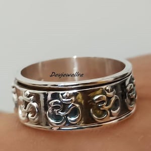 Könnte beinhalten: Ein silberner Drehring mit dem Om-Symbol, das um das Band graviert ist. Der Ring befindet sich an einem Finger.