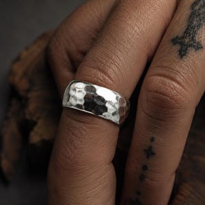 May include: A silver ring with a hammered texture. The ring is wide and has a slightly rounded shape. The ring is worn on a finger. The background is dark.