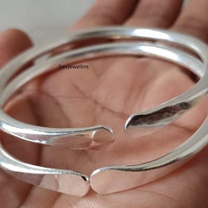 以下が含まれることがあります： 2つのオープンエンドのシルバーカラーのバングル。 ブレスレットは滑らかで磨かれた仕上げとわずかにハンマーで叩かれた質感を持っています。 デザインはシンプルでエレガントで、日常使いに適しています。 バングルは手の上に表示されています。