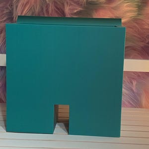 May include: A teal-colored, rectangular box with a cut-out section near the base. The box has a simple, modern design and is placed on a white slatted surface. The background features a fluffy, multi-colored texture.