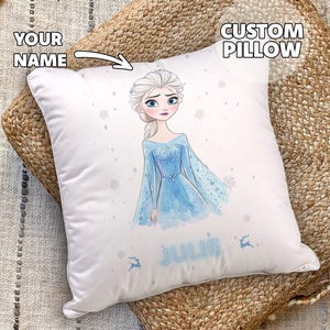Op de afbeelding: Een wit kussen met een aangepast ontwerp met een cartoonafbeelding van Elsa uit Frozen. Het kussen heeft de tekst "CUSTOM PILLOW" in de rechterbovenhoek en de tekst "JULIE" in het midden onderaan.
