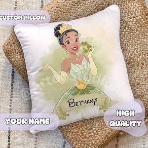 Op de afbeelding: Wit, op maat gemaakt kussen met een cartoonillustratie van een prinses die een kikker vasthoudt. De prinses draagt een groene jurk en een gouden kroon. Het kussen heeft de tekst "Bethany" op een banner. Het kussen is gelabeld "Custom Pillow", "Your Name" en "High Quality".