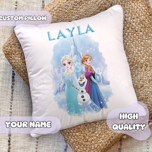 Op de afbeelding: Wit aangepast kussen met de naam "LAYLA" in blauw. Het kussen toont een kleurrijke illustratie van twee animatiefiguren en een sneeuwpop. De woorden "CUSTOM PILLOW", "YOUR NAME" en "HIGH QUALITY" zijn ook zichtbaar.