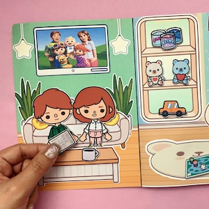 Könnte beinhalten: Eine farbenfrohe Papierpuppen-Szene, die ein Wohnzimmer mit Fernseher, Sofa und Couchtisch darstellt. Die Szene umfasst Papierpuppen eines Paares, einen Fernsehbildschirm mit einer Familie und dekorative Elemente wie ein Regal mit Spielzeug.