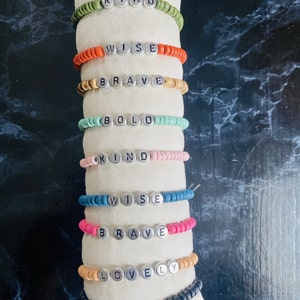Puede incluir: Una pila de pulseras de cuentas de colores con diferentes palabras positivas escritas en ellas, incluyendo "Happy", "Kind", "Wise", "Brave", "Bold", "Lovely".