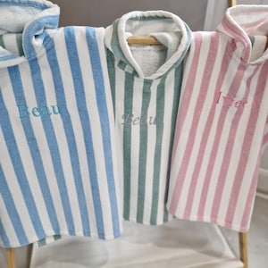 May include: Three striped hooded towels in blue, green, and pink, displayed on a wooden rack. Each towel features an embroidered name: "Beau" in blue, "Beau" in grey, and "Ivie" in pink. The towels are made of a soft, absorbent fabric, ideal for the beach or bath.