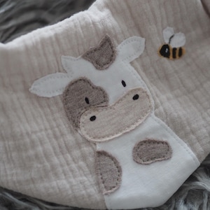Könnte beinhalten: Beige Baby-Lätzchen mit Kuh- und Bienenapplikation. Die Kuh ist weiß und braun, die Biene gelb und schwarz. Das Lätzchen ist aus weichem, strukturiertem Stoff gefertigt.