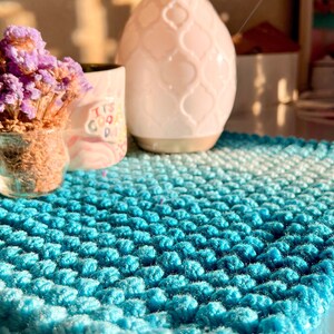 May include: A square, hand-crocheted coaster in shades of turquoise and white. The textured surface is made with a bobble stitch, creating a soft, plush appearance. The coaster is displayed with a small vase of dried purple flowers, a mug, and a decorative essential oil diffuser.