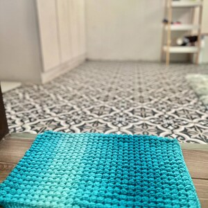 May include: A square, hand-crocheted rug in shades of turquoise and teal. The rug features a textured, bobble stitch pattern and is placed on a wooden surface. The background shows a patterned floor and a glimpse of a room.