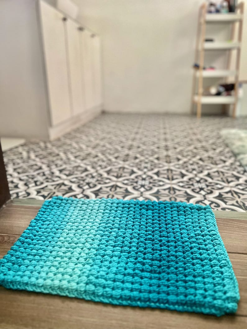 May include: A square, hand-crocheted rug in shades of turquoise and teal. The rug features a textured, bobble stitch pattern and is placed on a wooden surface. The background shows a patterned floor and a glimpse of a room.