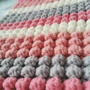 May include: Close-up of a handmade, textured blanket. The blanket features rows of pink, white, and gray bobbles, creating a soft, tactile surface. The color palette is a mix of pastel shades.