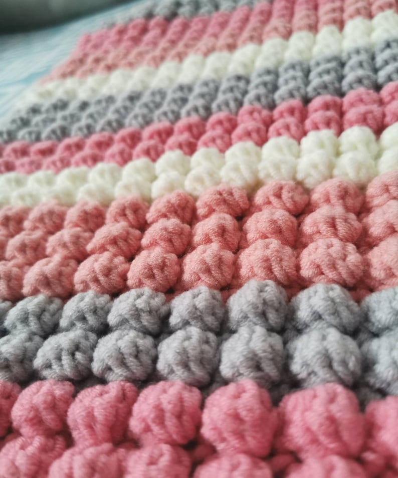 May include: Close-up of a handmade, textured blanket. The blanket features rows of pink, white, and gray bobbles, creating a soft, tactile surface. The color palette is a mix of pastel shades.