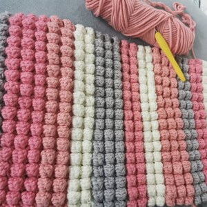 May include: A handmade crochet blanket in a striped pattern of pink, white, and gray. The blanket features a textured, bobble stitch design. A ball of pink yarn and a yellow crochet hook are visible in the background.