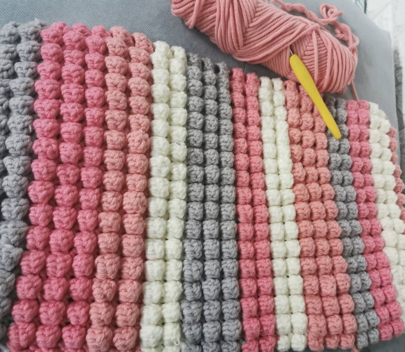 May include: A handmade crochet blanket in a striped pattern of pink, white, and gray. The blanket features a textured, bobble stitch design. A ball of pink yarn and a yellow crochet hook are visible in the background.
