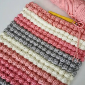 May include: A handmade crochet blanket in a diagonal striped pattern. The blanket features alternating rows of pink, white, gray, and a darker pink. A ball of pink yarn and a yellow crochet hook are in the upper right corner.