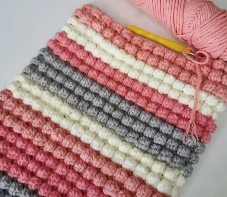 May include: A handmade crochet blanket in a diagonal striped pattern. The blanket features alternating rows of pink, white, gray, and a darker pink. A ball of pink yarn and a yellow crochet hook are in the upper right corner.